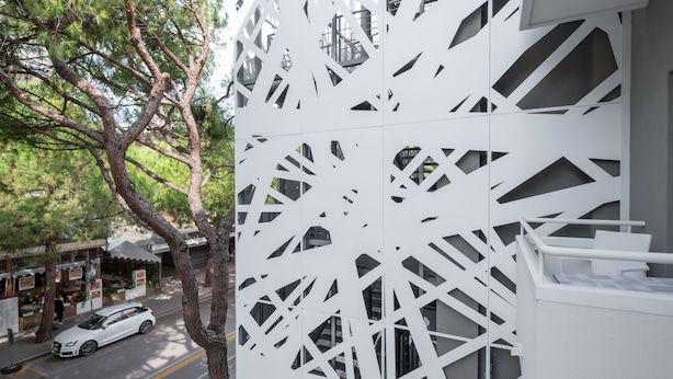 Um edifício moderno com padrões geométricos recortados na fachada, um carro branco estacionado na rua e uma árvore em primeiro plano.