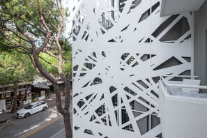 A modern building with geometric cut-out patterns on its facade, a white car parked on the street, and a tree in the foreground.