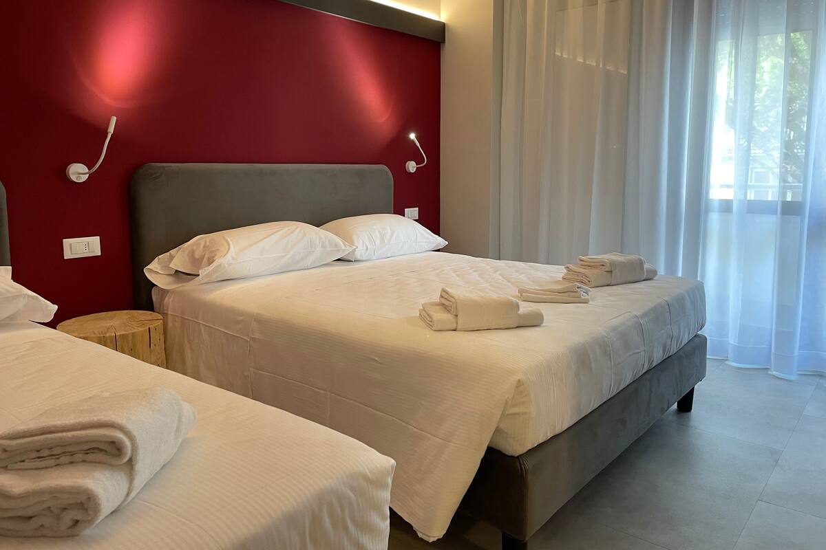 A hotel room with two beds, white linens, a red accent wall, and a large window with sheer curtains.