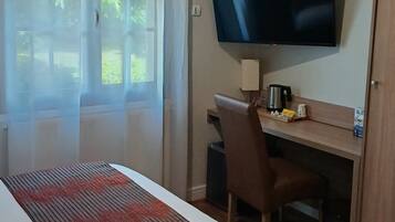 A hotel room with a bed, a desk with a chair, a television, and a window with sheer curtains.