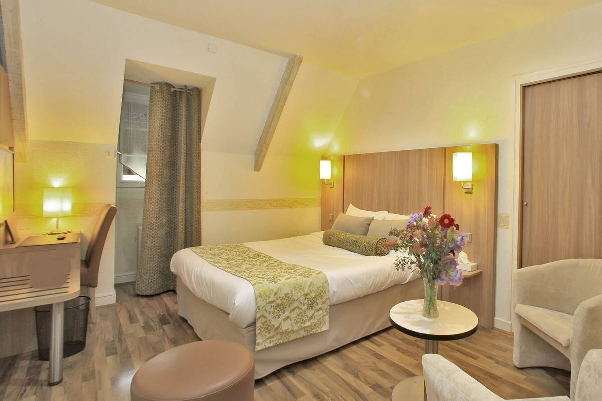 A hotel room with a bed, bedside table, chair, and a small round table with a vase of flowers.