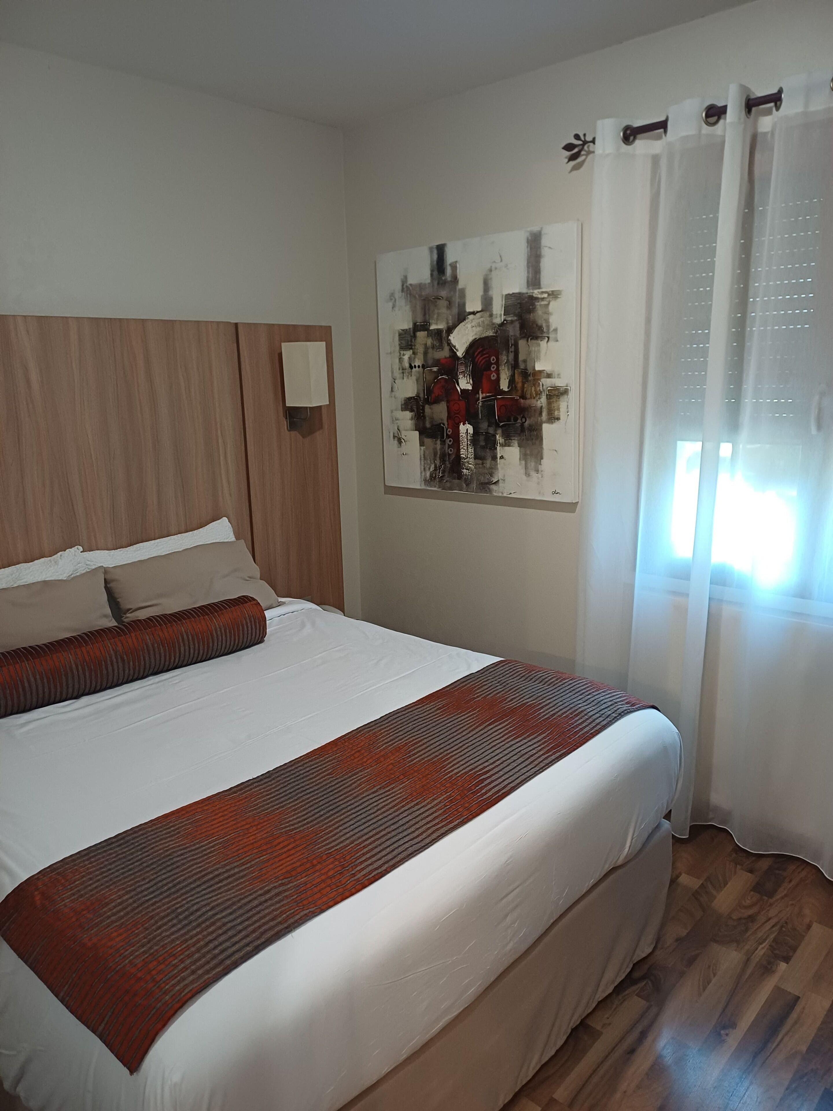 Una cama bien hecha con una colcha estampada, un cabecero de madera, una obra de arte en la pared y una ventana con cortinas traslúcidas.