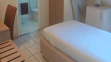 Quarto com cama, uma cadeira de madeira, uma mesa pequena, guarda-roupa e banheiro visível por uma porta aberta.