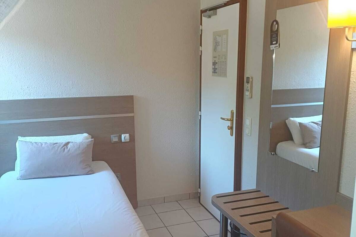 A hotel room with a bed, a desk, a chair, a mirror, and a door.