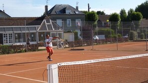 A tennis court with a net, a player in red shorts, and a house in the background.