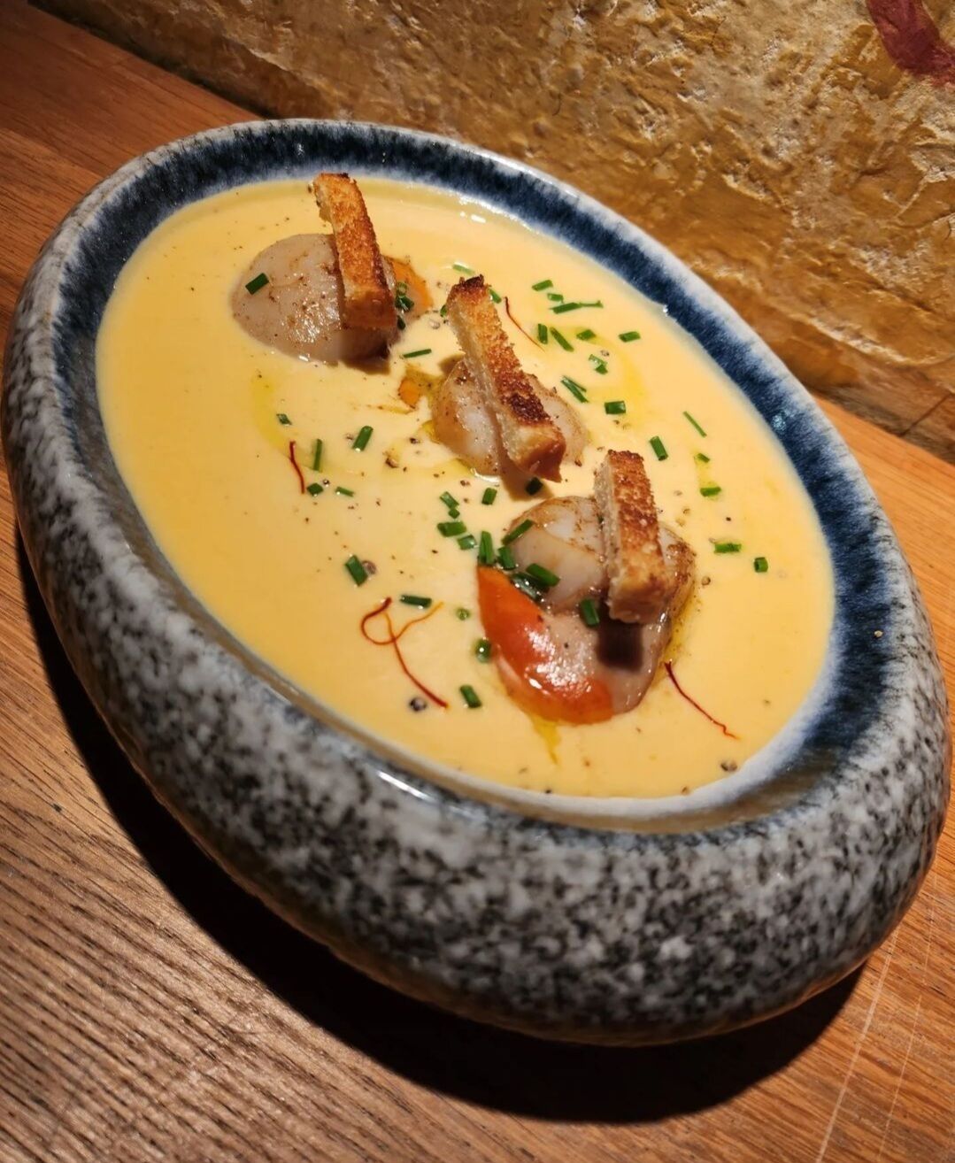 Un tazón de sopa cremosa con pan y cebollines sobre una mesa de madera.