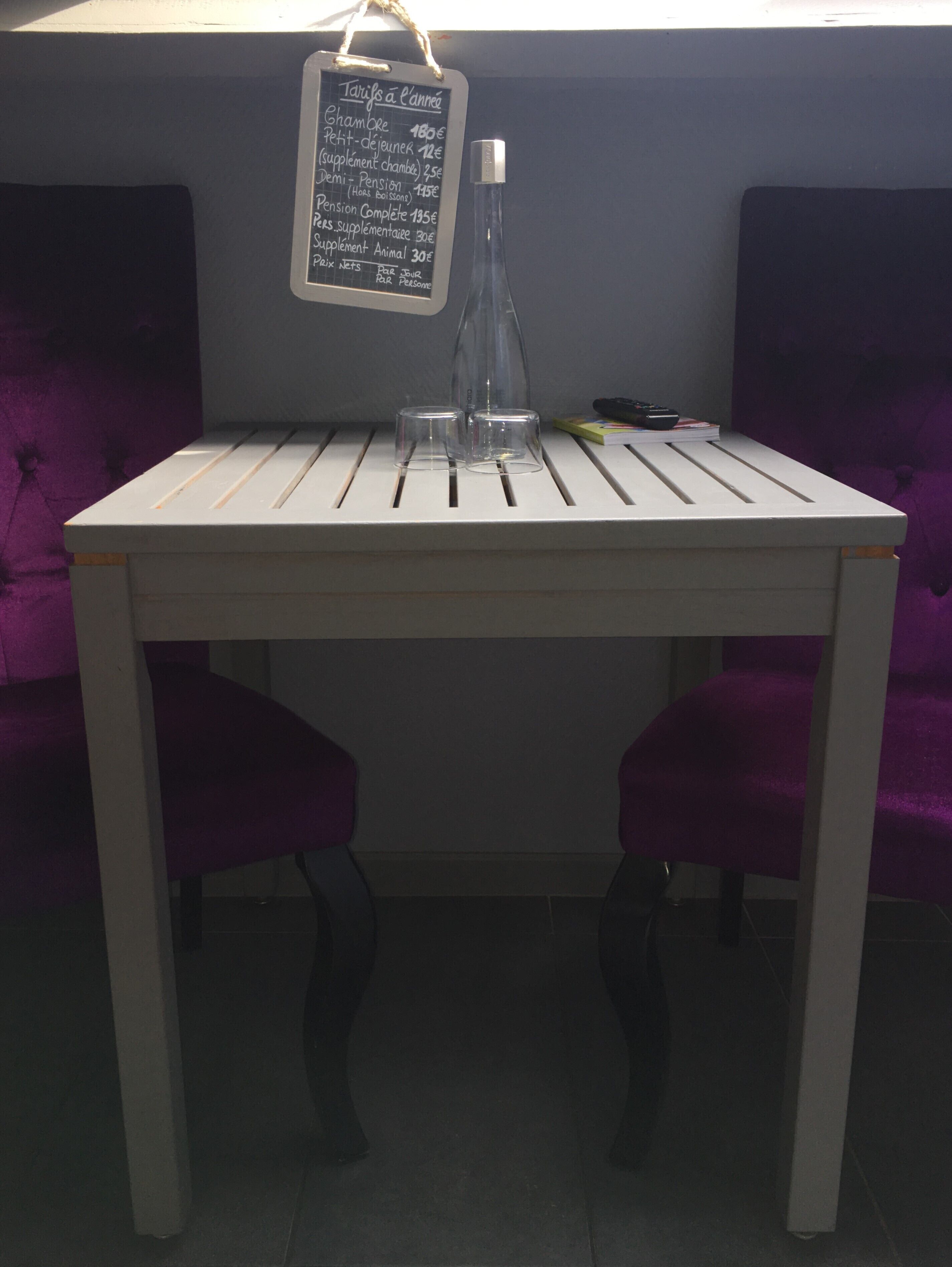 A small white table with two purple upholstered chairs, a glass bottle, and a chalkboard menu listing prices for various items.