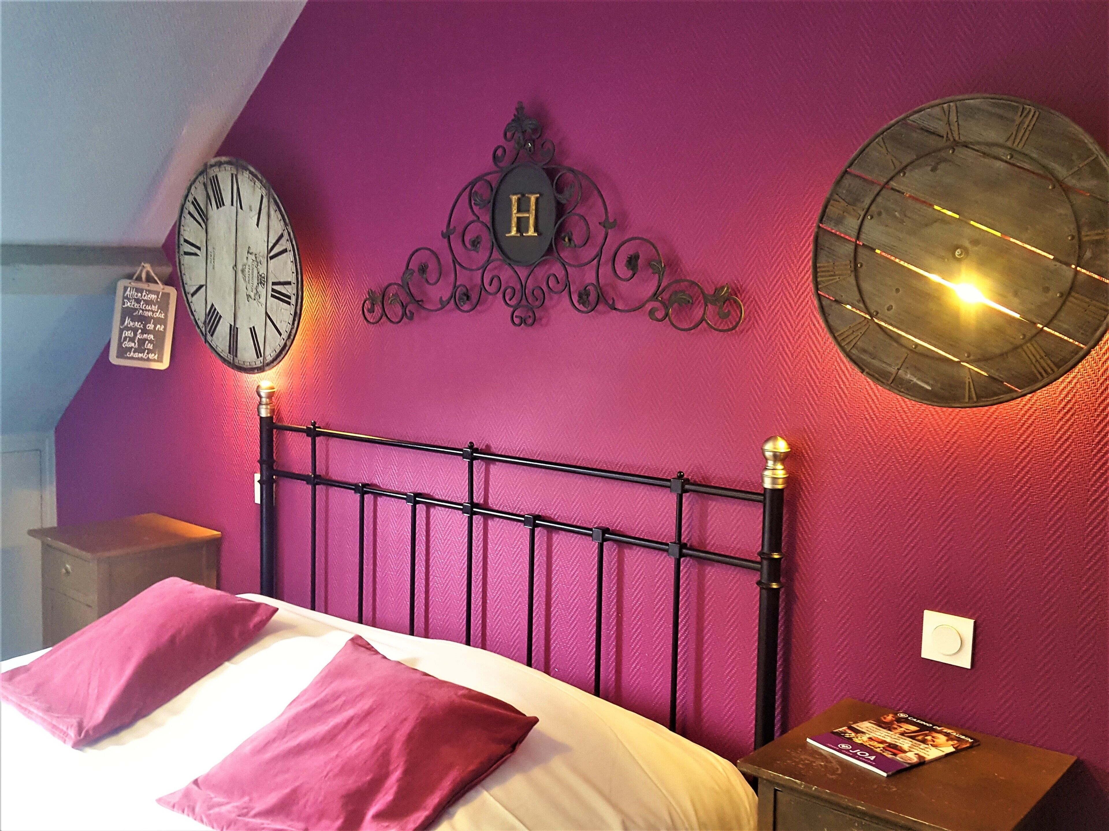 A bedroom with a bed, two pillows, a nightstand, a clock, a decorative letter 'H', and a wall-mounted light fixture.