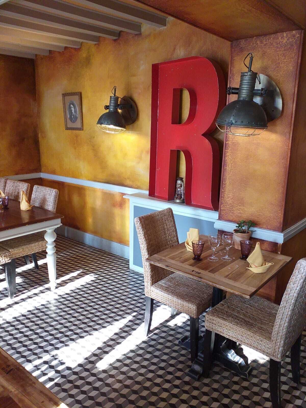 A restaurant with a checkered floor, wicker chairs, wooden tables, a large red letter 'R' on the wall, and pendant lights.