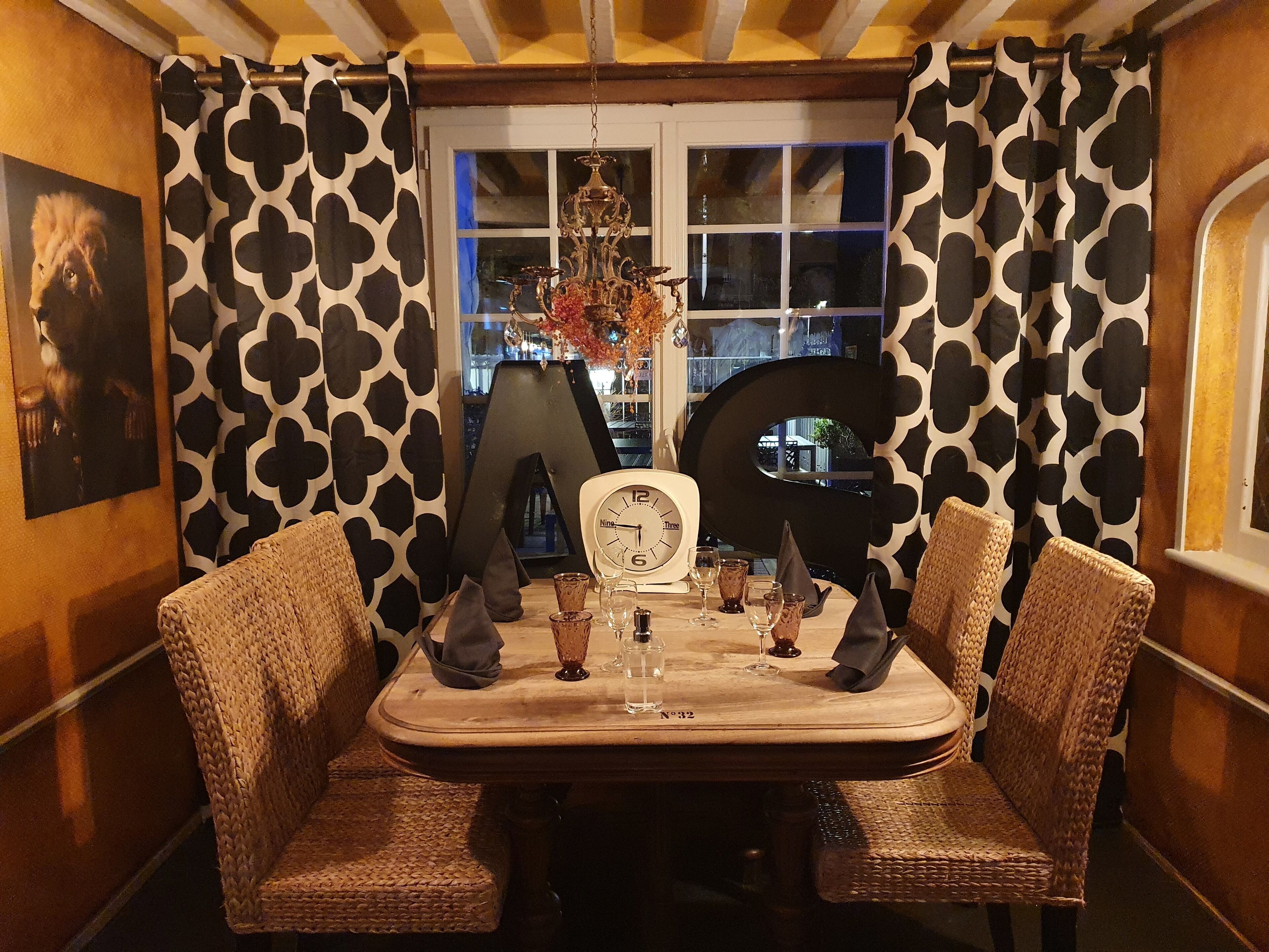 Un comedor con una mesa lista para dos personas, que incluye un reloj, vasos y un jarrón con flores.