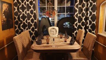 Un comedor con una mesa lista para dos personas, que incluye un reloj, vasos y un jarrón con flores.