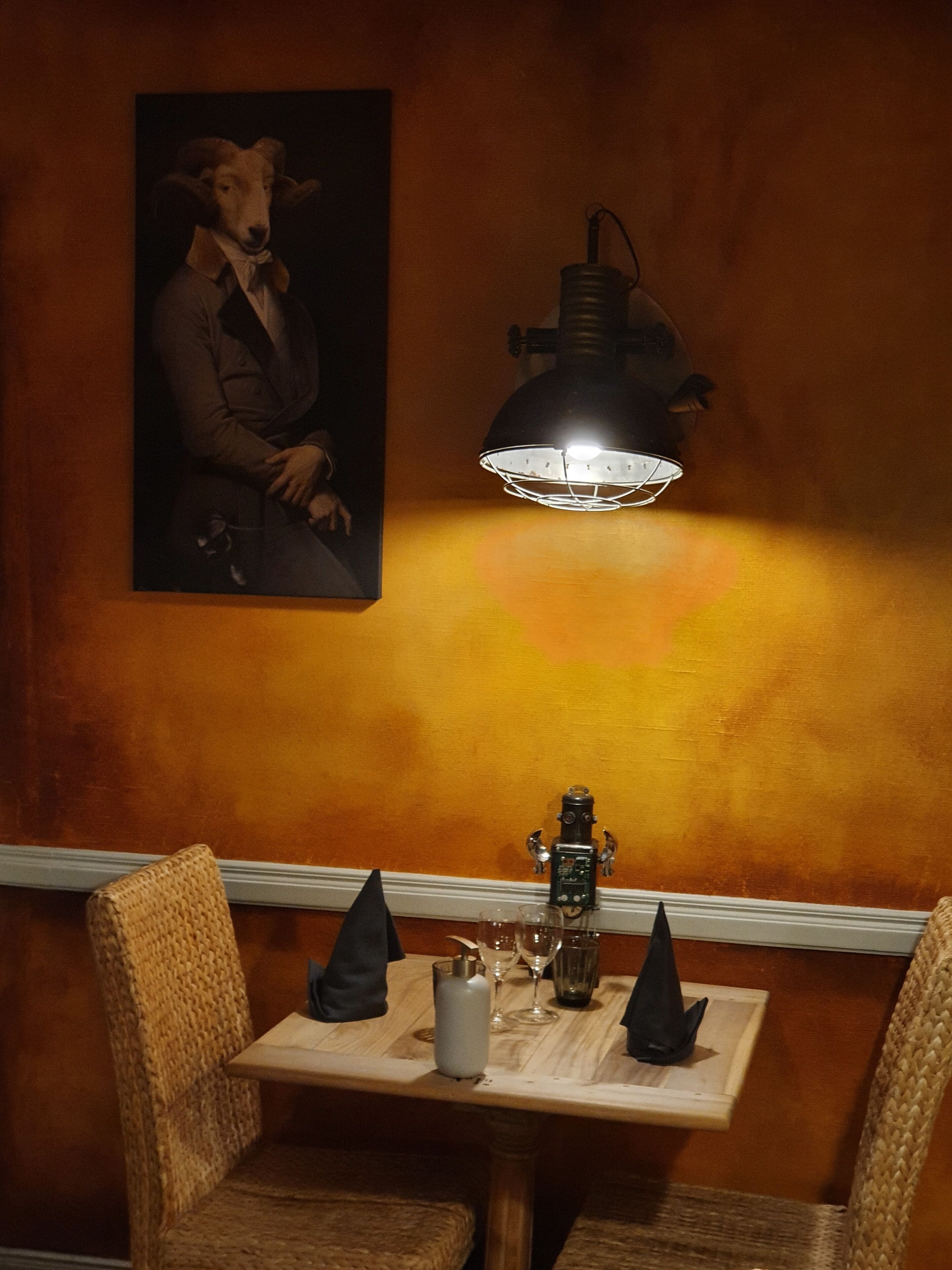 A dining area with a table set for two, a wall-mounted lamp, and a framed portrait above the table.