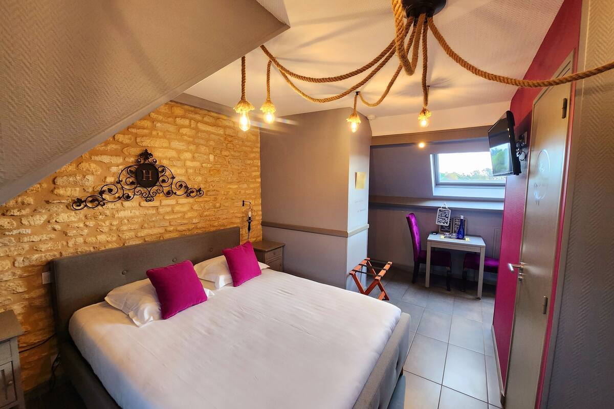 A bedroom with a large bed, a wooden headboard, a brick wall, a chandelier, a desk, and a chair.