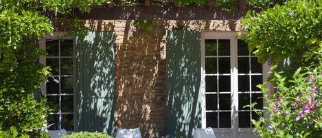 Ein überdachter Patio-Bereich mit zwei weißen Stühlen, einer Steinmauer und einem Fenster mit grünen Fensterläden.