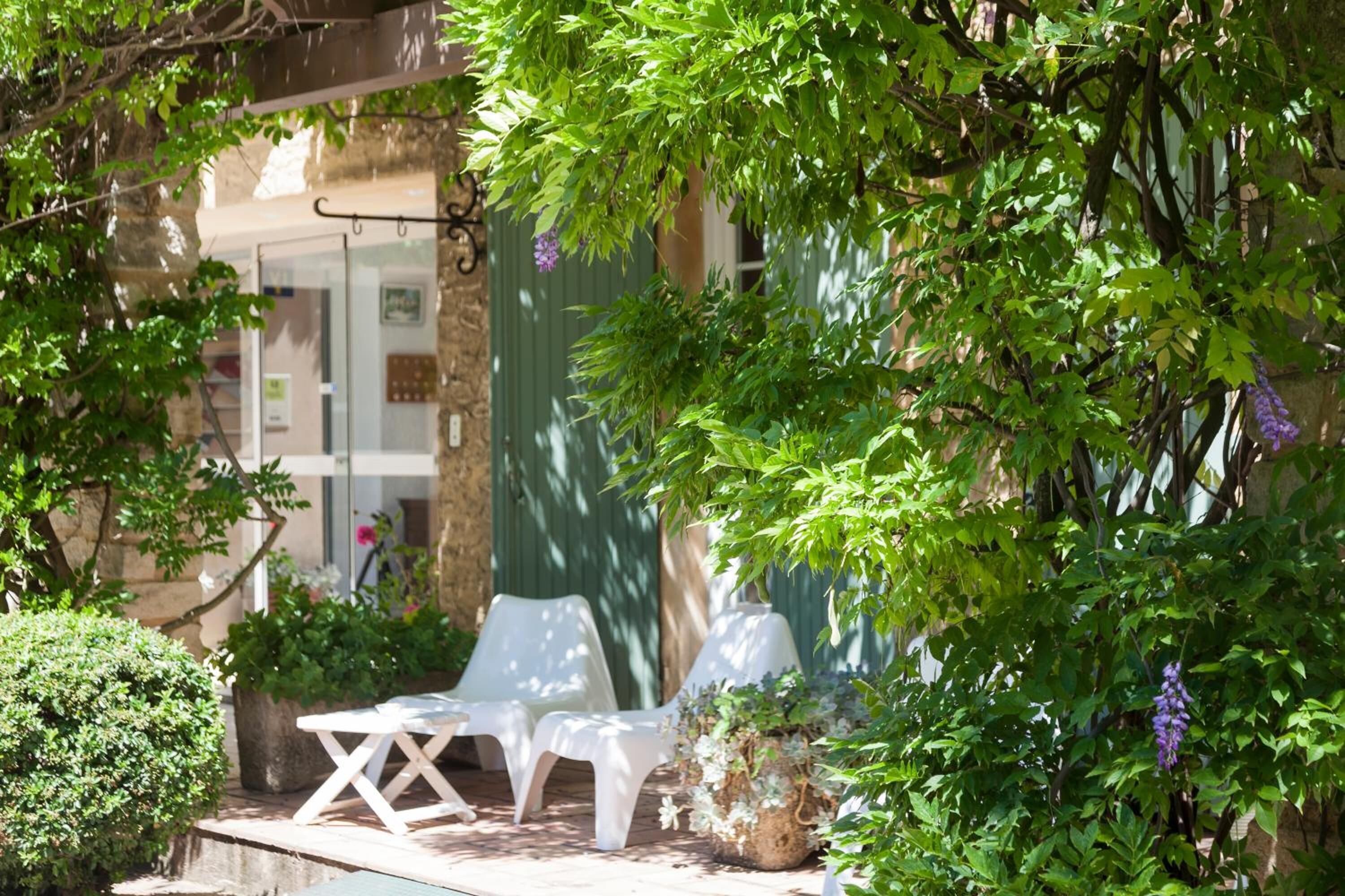 A patio area with white chairs and tables, surrounded by lush greenery and a pergola.