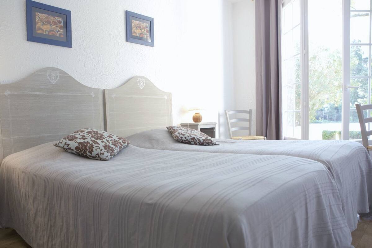A double bed with floral pillows, a nightstand with a lamp, and a chair by a window with curtains.