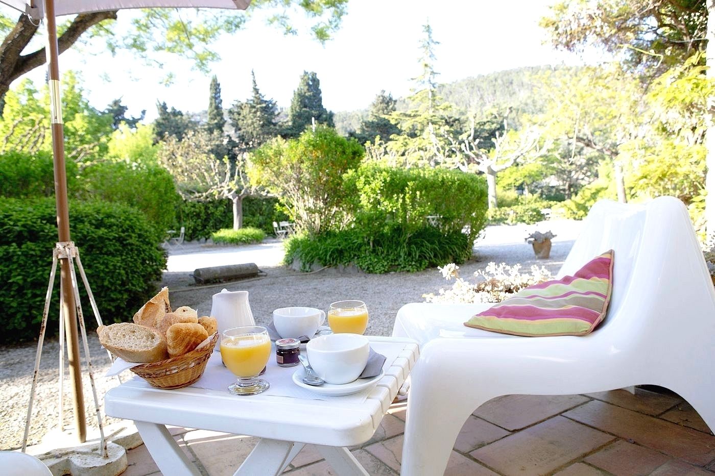 A breakfast setup with bread, juice, and coffee on a table outdoors, with a chair and a view of greenery and hills in the background.