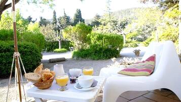 Un desayuno con pan, jugo y café sobre una mesa al aire libre, con una silla y vistas a zonas verdes y colinas al fondo.