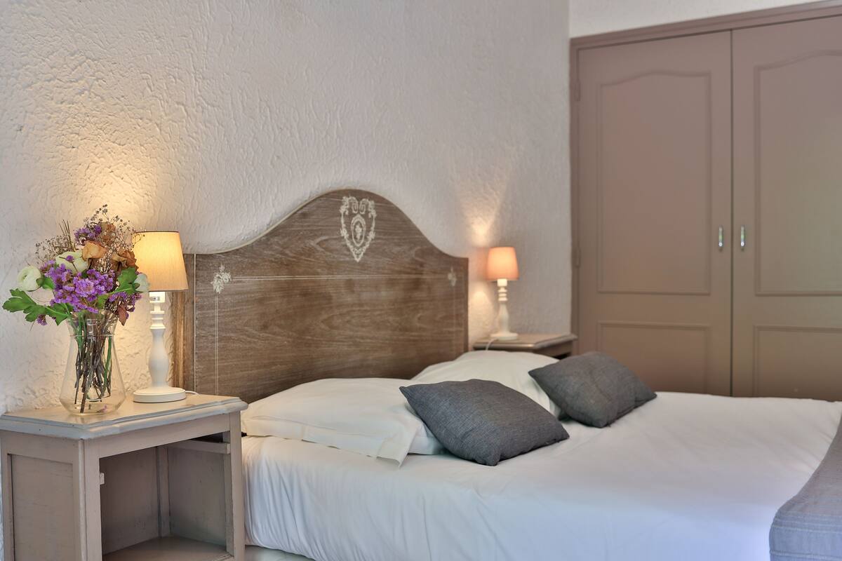 A neatly made bed with white linens and two pillows, a wooden headboard with a heart design, a bedside table with a lamp, and a vase with flowers.