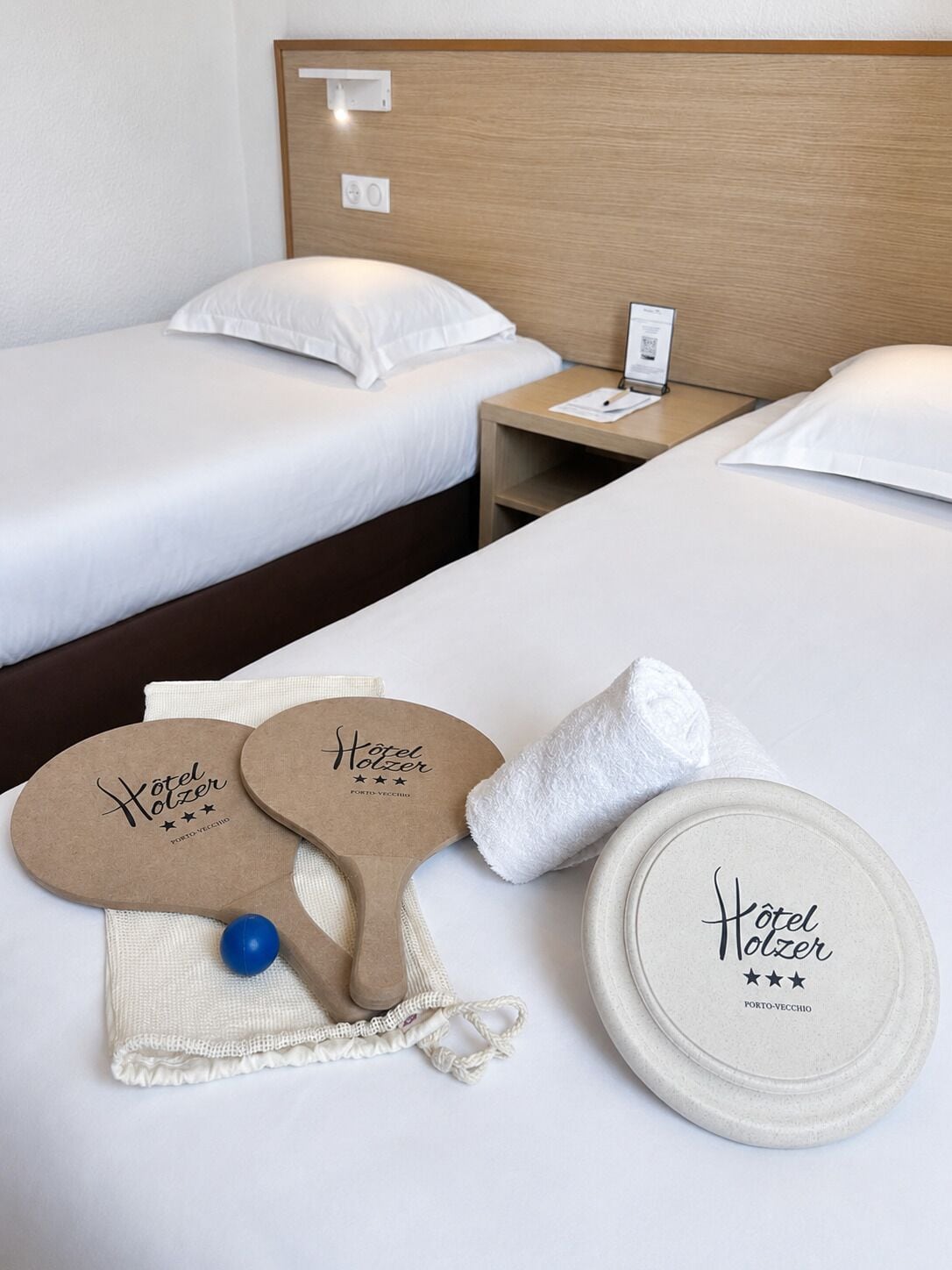 Habitación de hotel con dos camas, una mesita de noche y comodidades que incluyen una toalla, un tapete de baño y una bandeja redonda con el nombre del hotel.