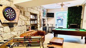A room with a foosball table, dartboard, and pool table, featuring stone walls and a green wall with plants. - Hôtel Holzer II (Porto-Vecchio)