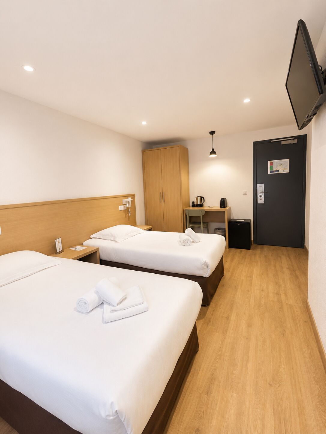 A hotel room with two beds, a wooden wardrobe, a desk with a chair, a television mounted on the wall, and a door with a lock.
