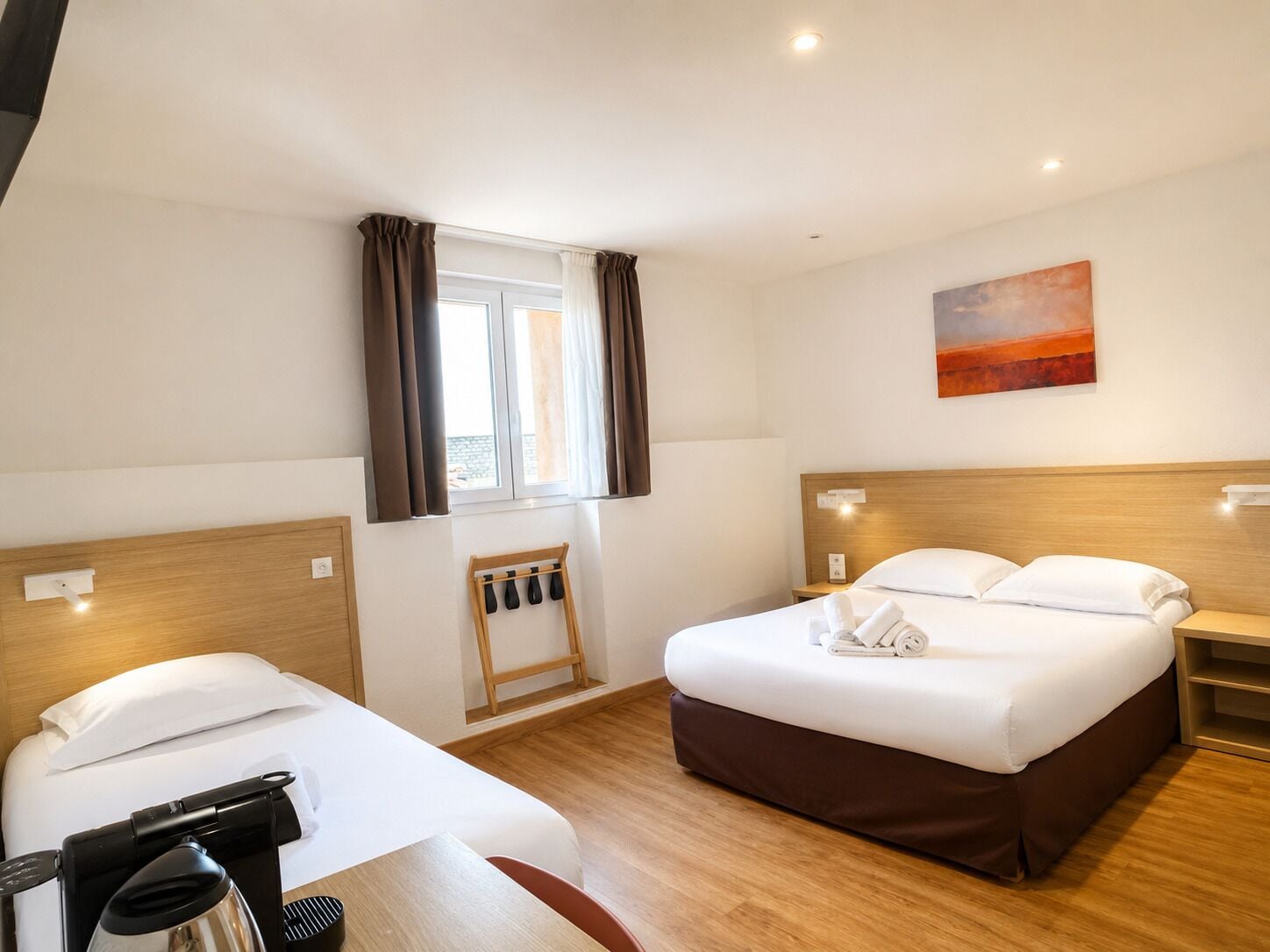 Una camera d'albergo con un letto, una scrivania con una macchina del caffè, una finestra con tende e un quadro appeso al muro.
