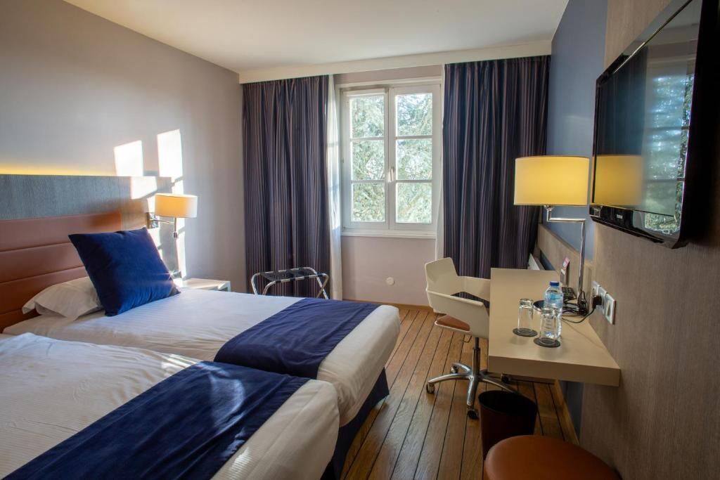 A hotel room with two beds, a desk with a chair, a TV, and a window with curtains.