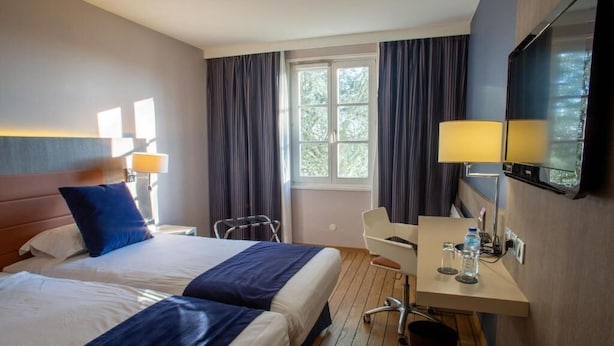A hotel room with two beds, a desk with a chair, a TV, and a window with curtains. - Les Tuileries (Fey)