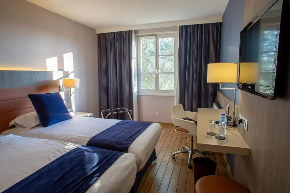 A hotel room with two beds, a desk with a chair, a TV, and a window with curtains.