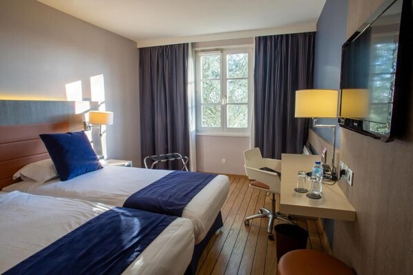 Habitación de hotel con dos camas, un escritorio con silla, televisor y ventana con cortinas.