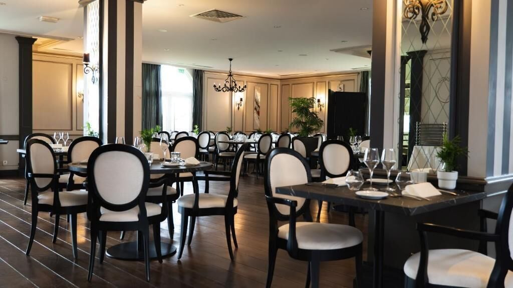 Un comedor amplio con varias mesas y sillas, ventanas grandes y una araña.
