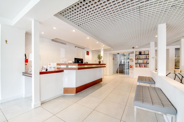 A modern reception area with a wooden reception desk, a seating area with benches, and a bookshelf filled with books. - Brit Hotel Porte d’Espagne (Perpignan)