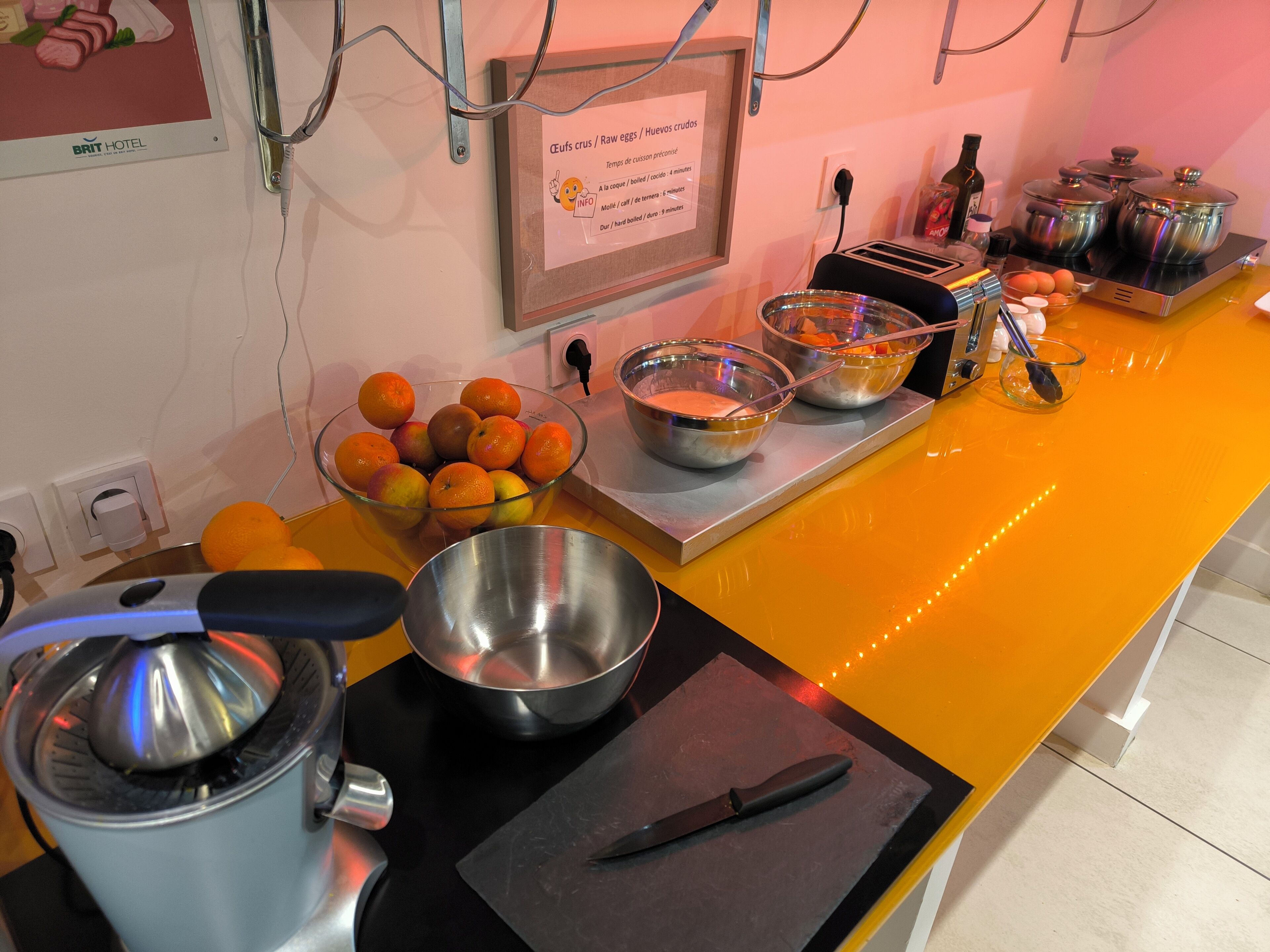 Una cocina con una encimera que muestra diversos utensilios de cocina, cuencos y un exprimidor. Hay naranjas y un letrero con instrucciones de cocina.