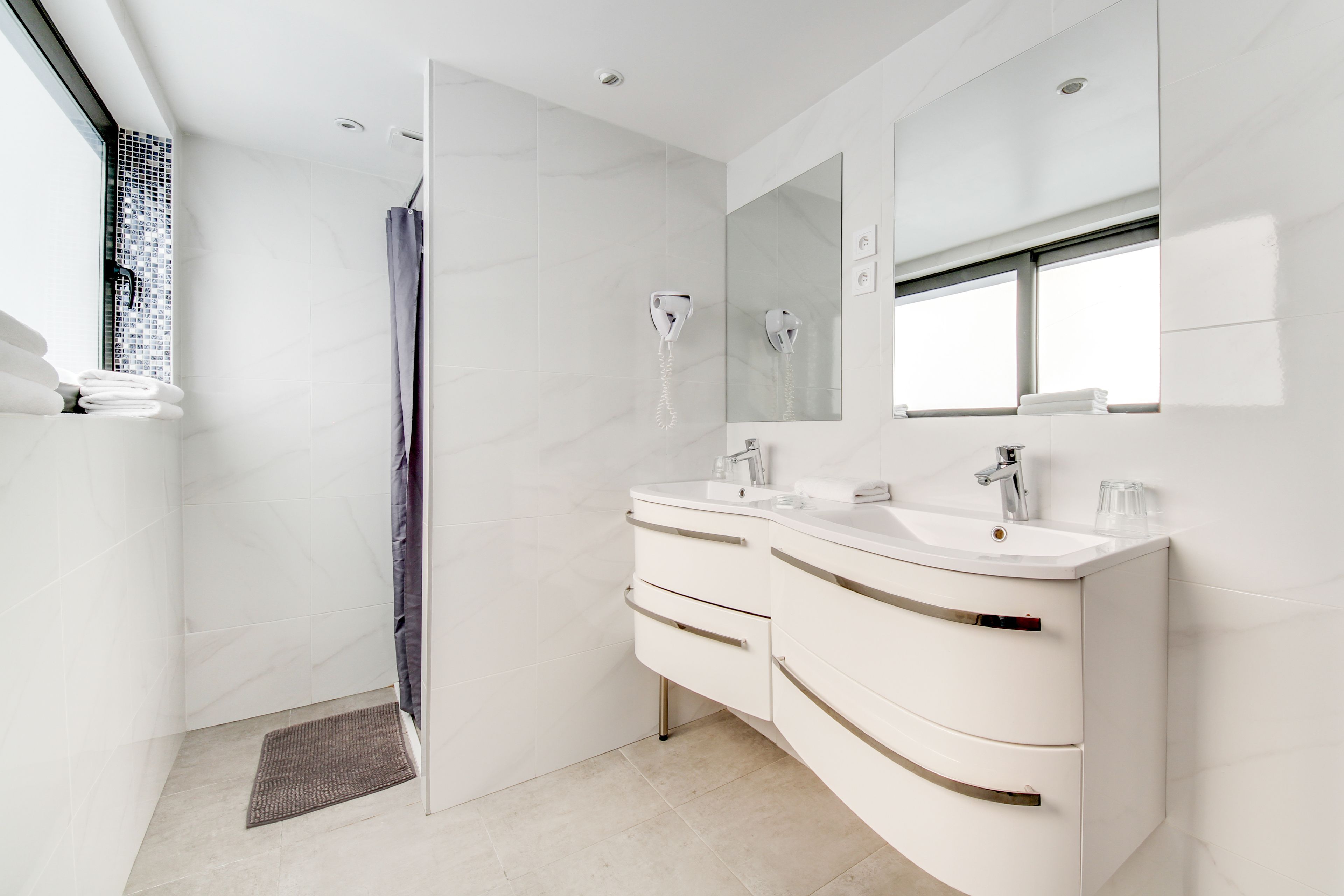 A modern bathroom with a walk-in shower, two sinks, a large mirror, and a window.