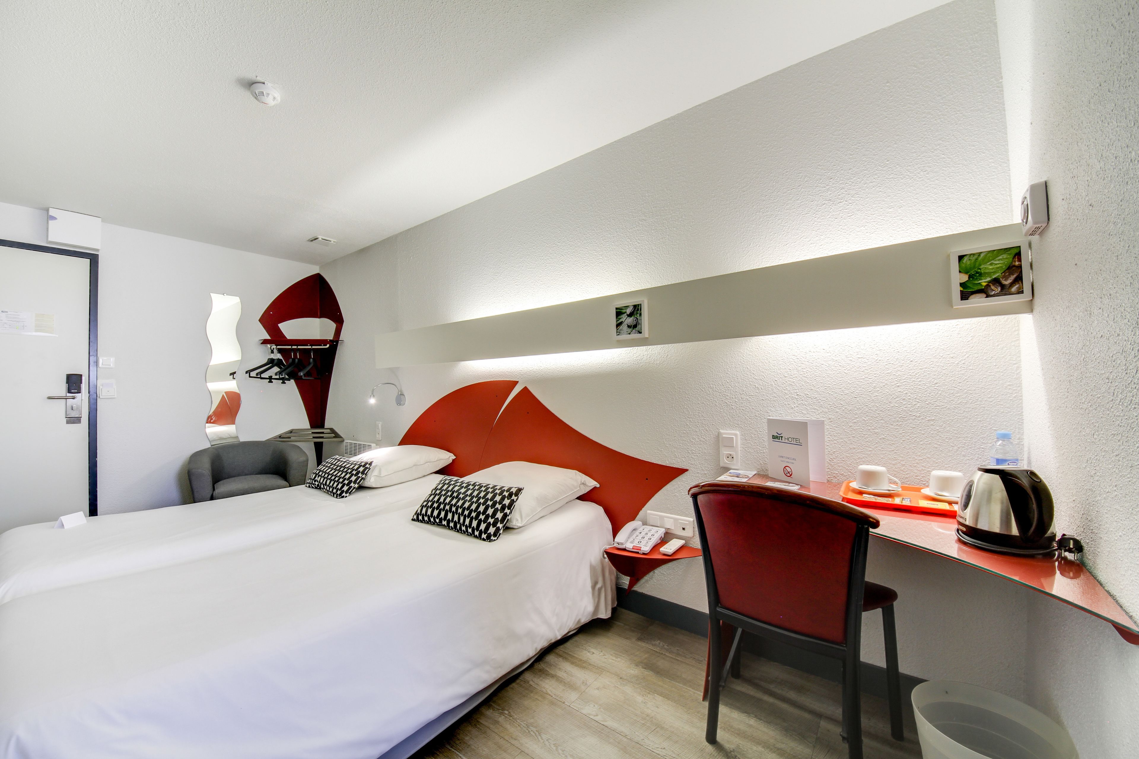 A hotel room with a bed, a desk, a chair, a telephone, and a small shelf with a plant and a kettle.