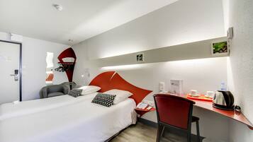 A hotel room with a bed, a desk, a chair, a telephone, and a small shelf with a plant and a kettle.