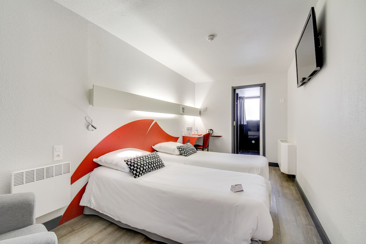 A hotel room with two beds, a desk, a chair, a TV, and a door.