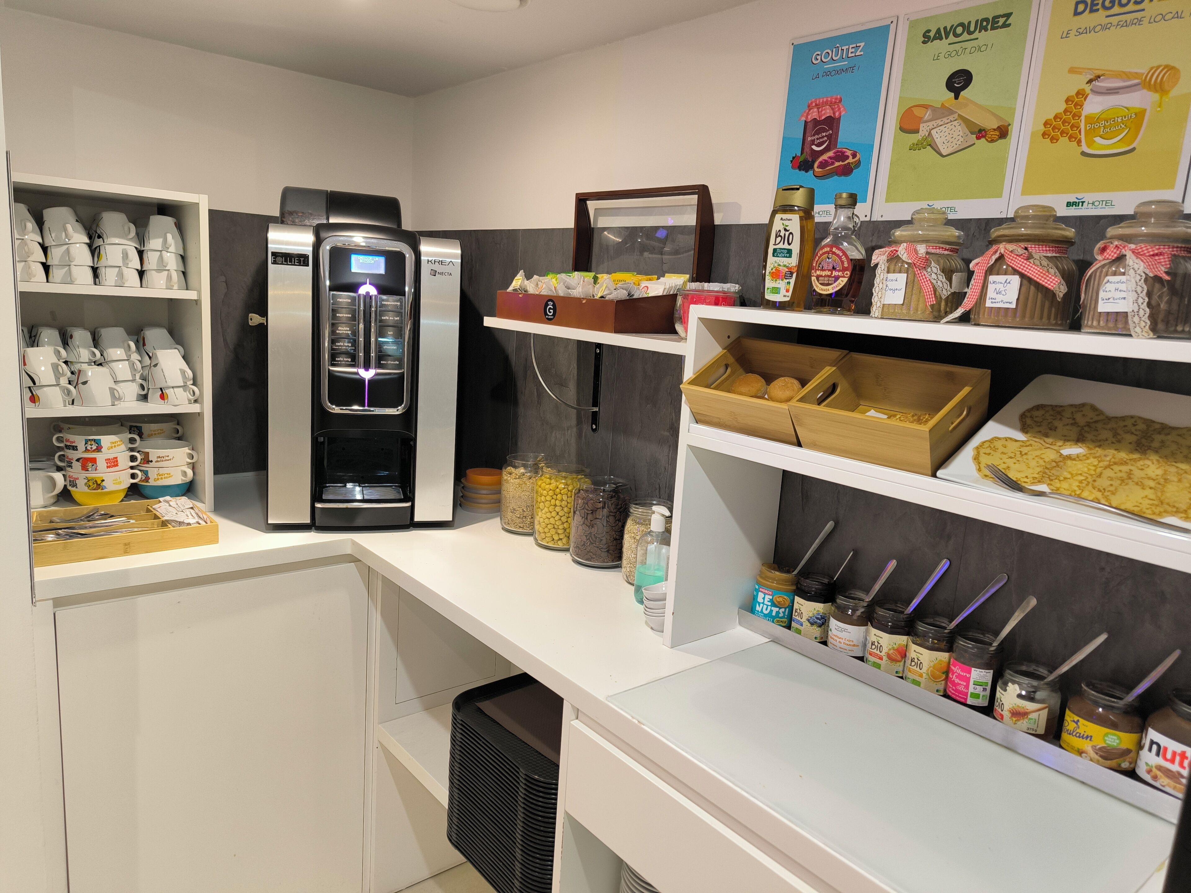 A hotel kitchen area with a coffee machine, shelves with jars of condiments, and a display of mugs.