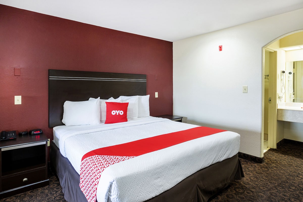 A hotel room with a large bed, a red pillow, a bedside table with a phone, and a mirror in the corner.