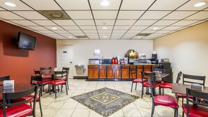 A hotel dining area with red chairs, a tiled floor, a kitchen counter, and a flat-screen TV mounted on the wall. - OYO Hotel LSU (Port Allen)
