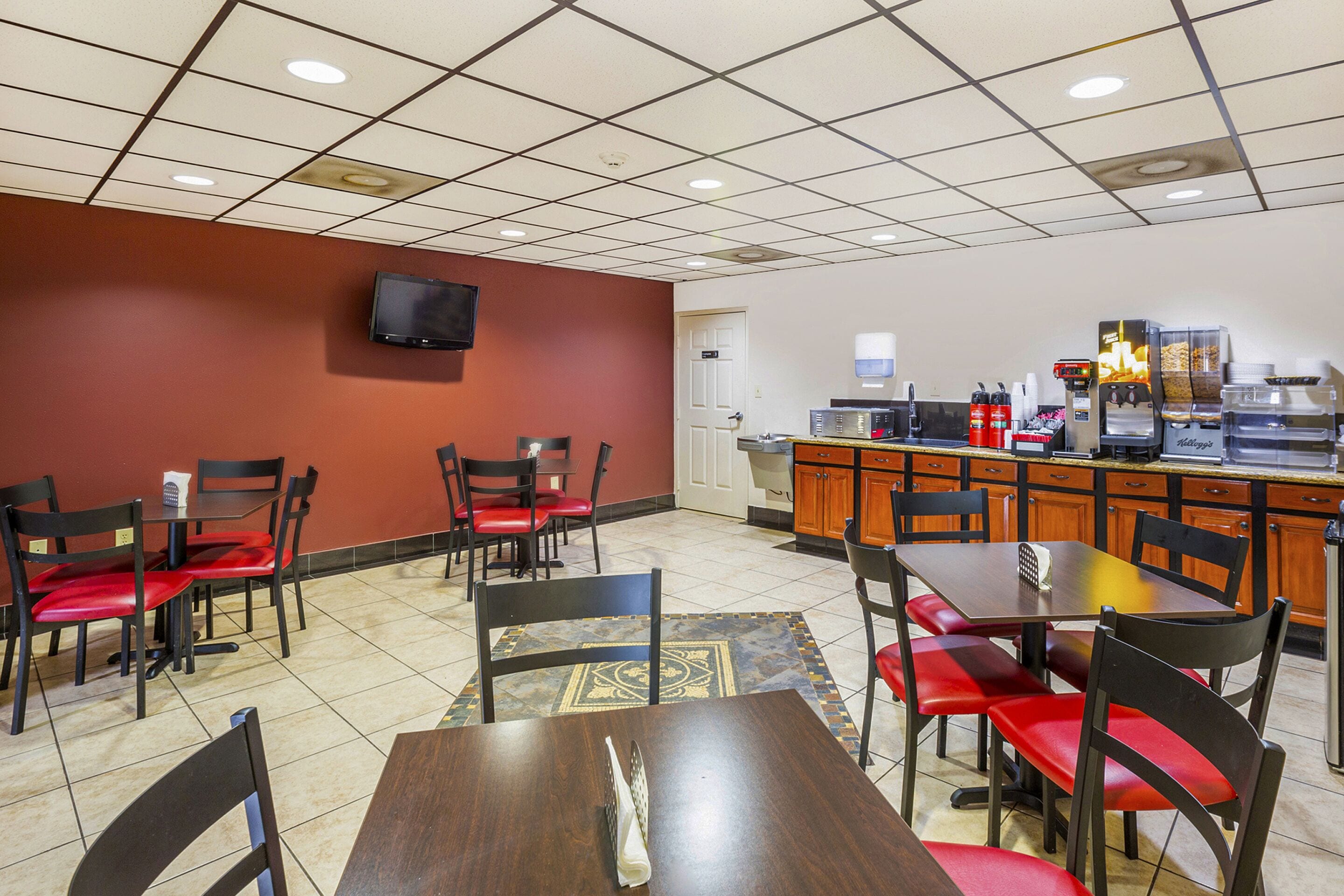 A restaurant with a tiled floor, red and black chairs, a wooden table, a television mounted on the wall, and a kitchen area with a coffee machine and condiment dispensers.