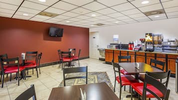 A restaurant with a tiled floor, red and black chairs, a wooden table, a television mounted on the wall, and a kitchen area with a coffee machine and condiment dispensers.