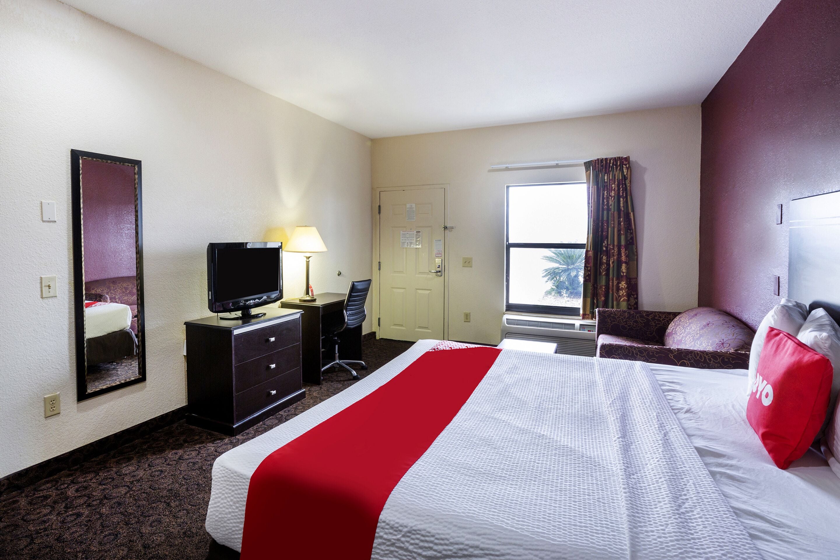 A hotel room with a bed, a TV on a dresser, a desk, and a window with curtains.