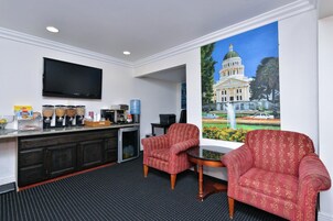 Habitación de hotel con kitchenette, televisor de pantalla plana, dos sillones estampados, zona de café y un amplio ventanal con vistas a un edificio.
