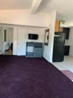 Amplia sala de estar con un gran tapete oscuro, una cocina con refrigerador y armarios, y un televisor en la pared.