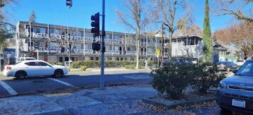 Un edificio de varios pisos con un diseño arquitectónico moderno, un semáforo y un estacionamiento con coches.