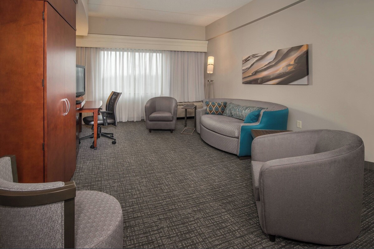A hotel room with a grey sofa, a grey armchair, a wooden cabinet, a desk with a chair, a flat-screen TV, and a large window with curtains.