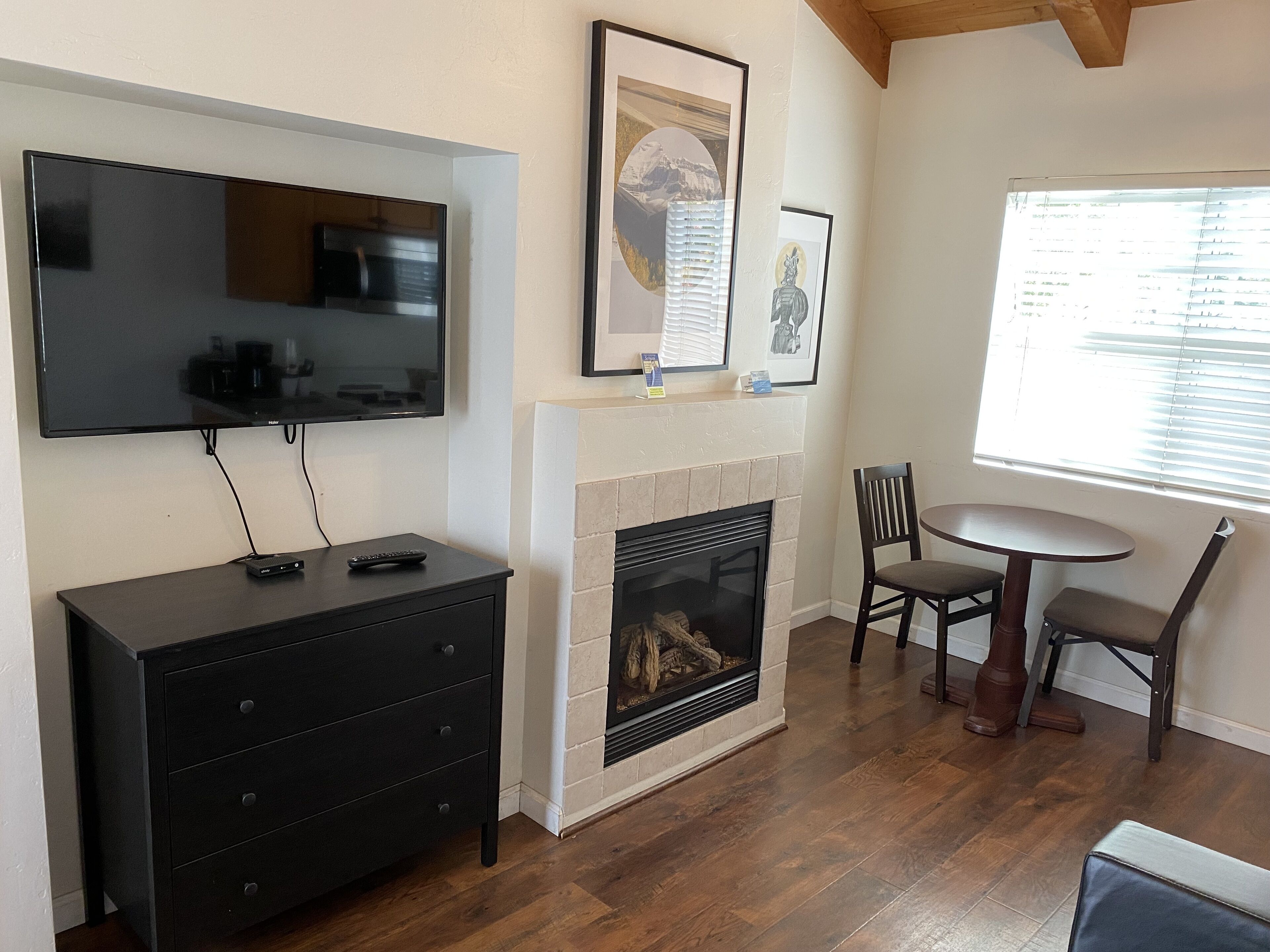 A living room with a fireplace, a TV mounted on the wall, a dining table with chairs, and framed artwork on the wall.