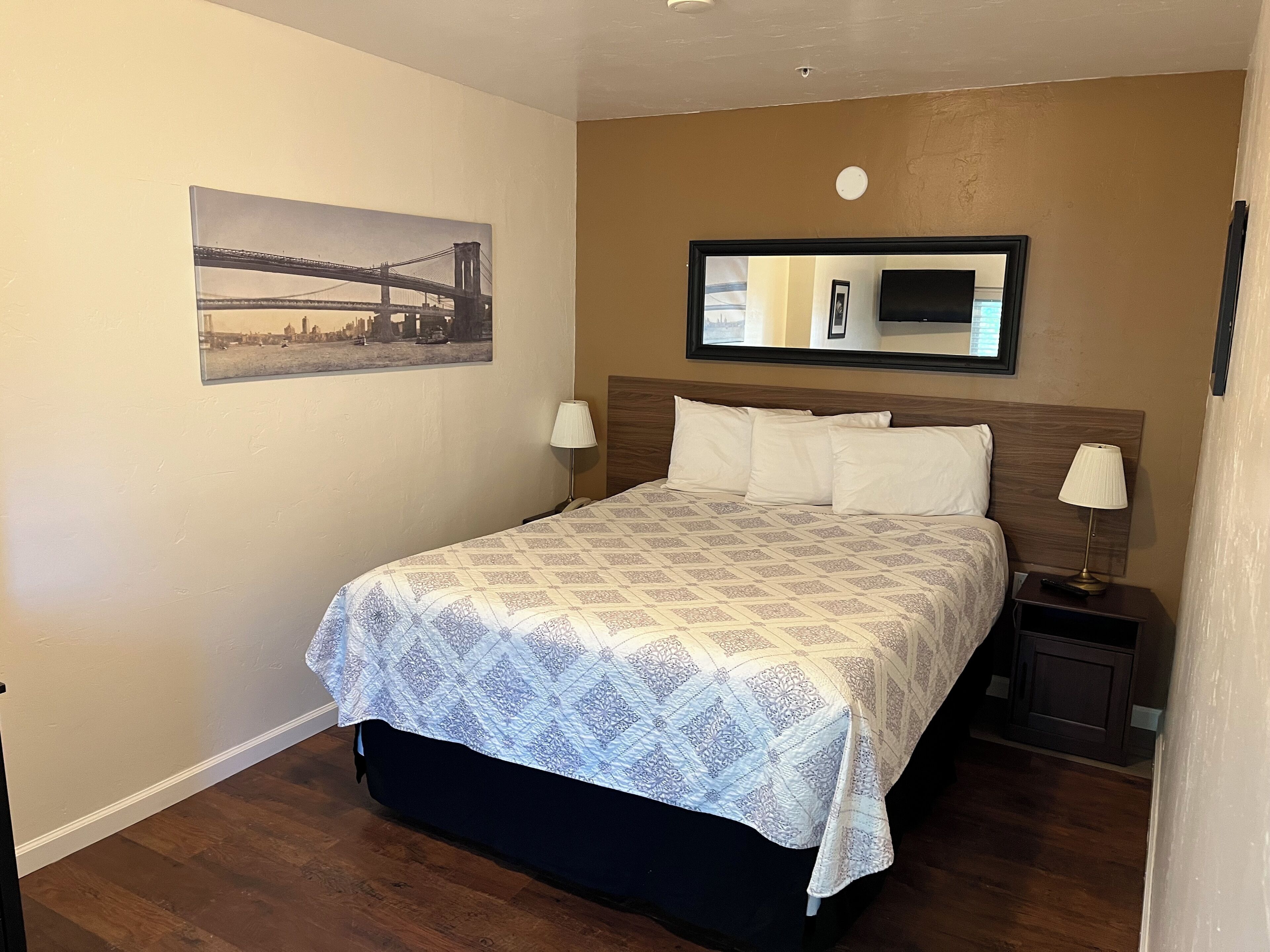 A hotel room with a bed, two bedside lamps, a TV on the wall, and a framed picture of a bridge.